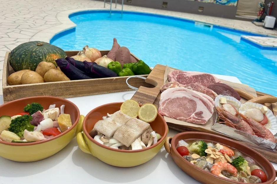 プールサイドで準備された、豪華なバーベキュー食材のセットです。牛肉、豚肉、エビ、ホタテなどの魚介類、そしてカボチャやナス、ジャガイモなどの新鮮な野菜が豊富に並んでいます。