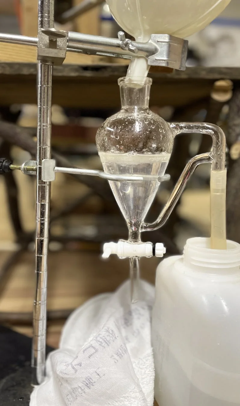 実験器具を用いた液体の分離作業の様子。分液ロートで液体が分離され、下の栓から別の容器に排出されている。化学実験の一場面。