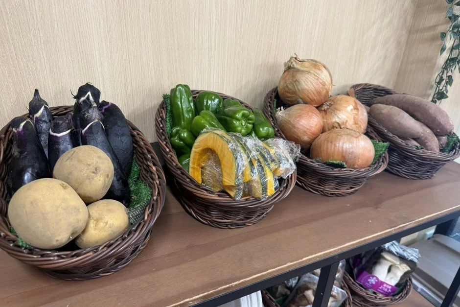木製の棚に、なす、じゃがいも、ピーマン、かぼちゃ、玉ねぎ、さつまいもなどの新鮮な野菜が籠に美しく陳列されている。下段にはきのこ類も見え、産直販売所やスーパーの青果コーナーのようだ。