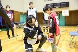 体育館で、ゼッケンを着用した大人が子供と向き合い、しゃがんで何らかの測定のような活動をしている。