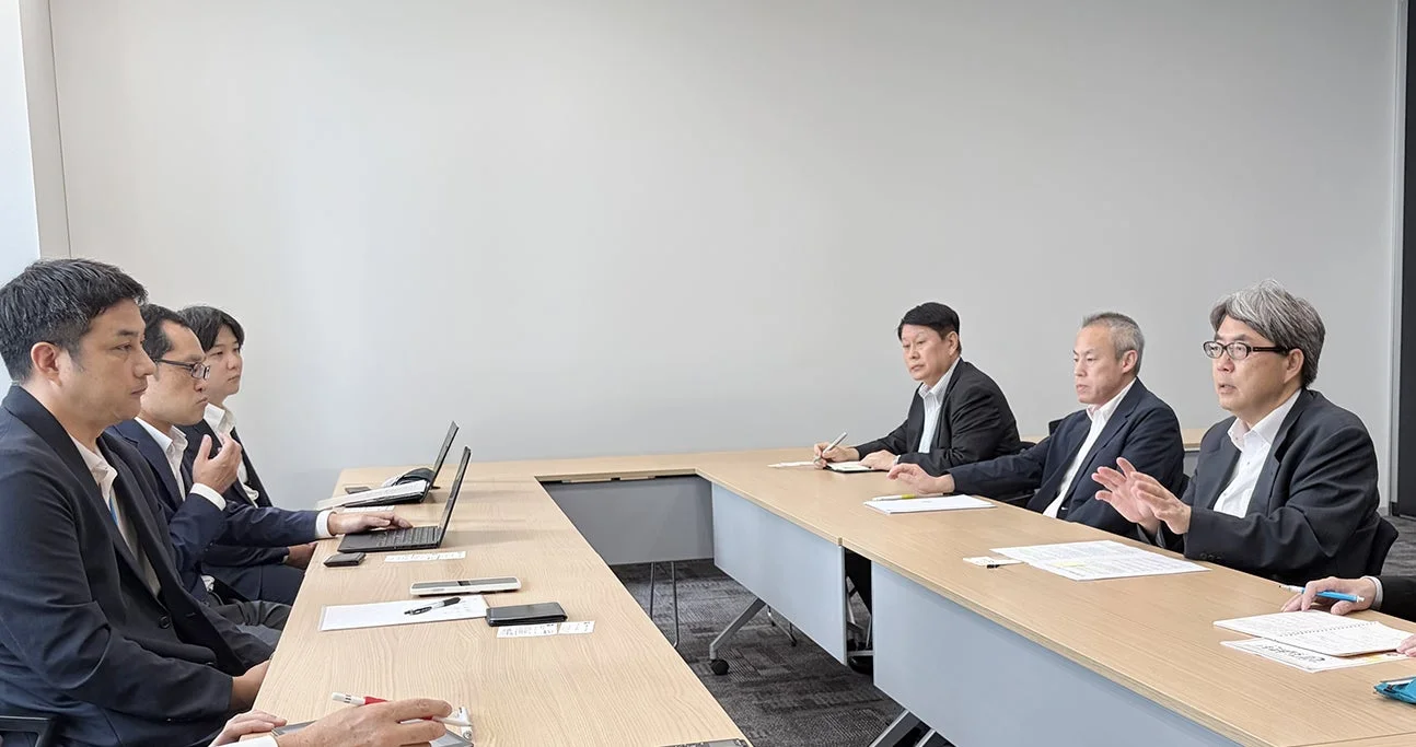 複数のビジネスマンが会議室のテーブルを囲んで座り、ミーティングを行っている様子です。一人が話しており、他のメンバーは聞き入ったり、資料を見たりしています。