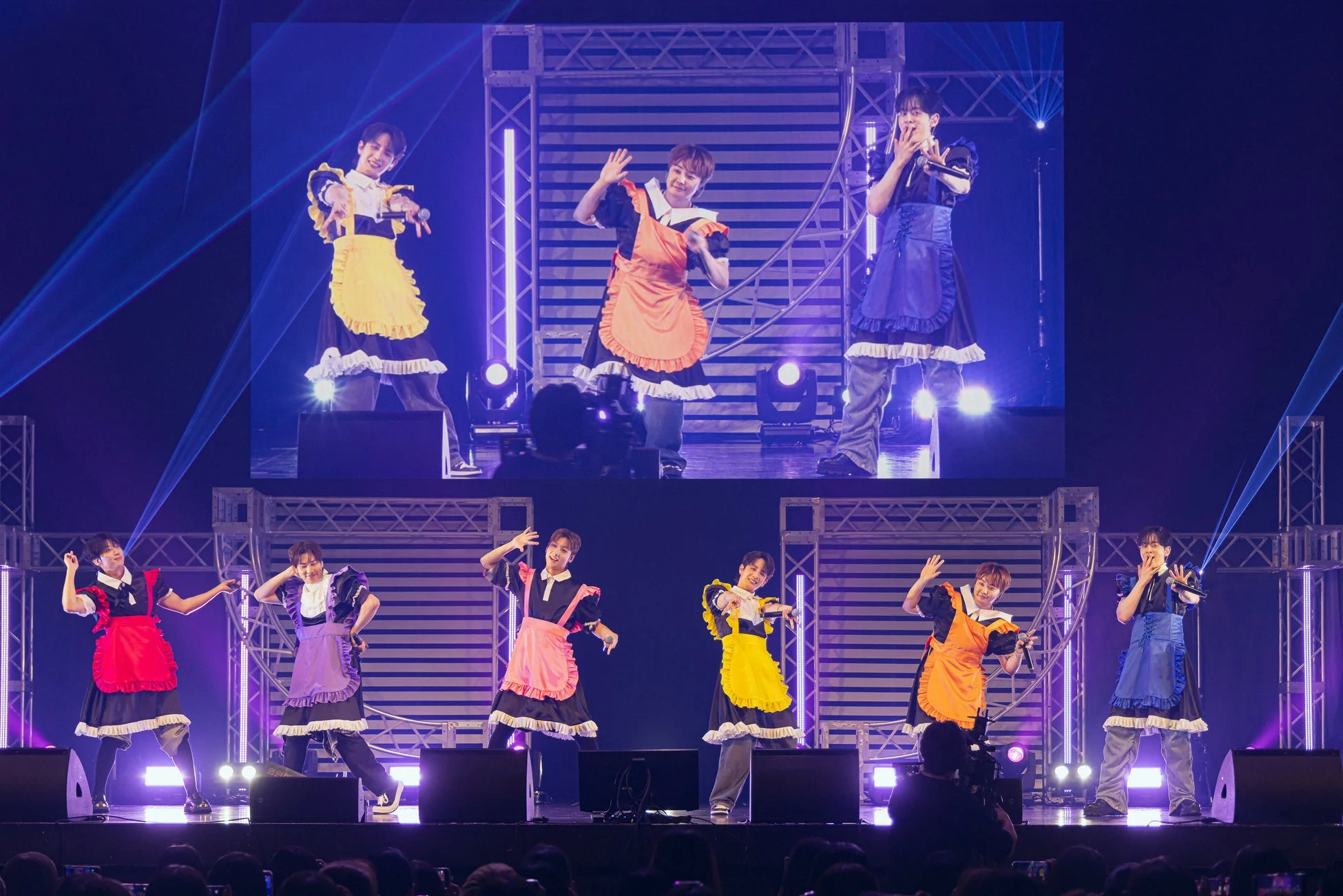 カラフルなエプロンのメイド服を着用した男性アイドルグループが、照明が輝くステージでパフォーマンスをしている様子。背後の大型スクリーンにはメンバーのクローズアップが映し出されています。