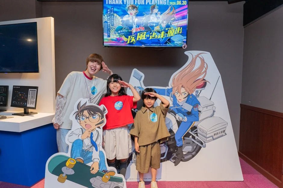 名探偵コナンとリアル脱出ゲームのコラボイベント会場で、大人と子供2人がコナンと警察官のキャラクターパネルと一緒に敬礼ポーズで記念撮影をしている様子です。