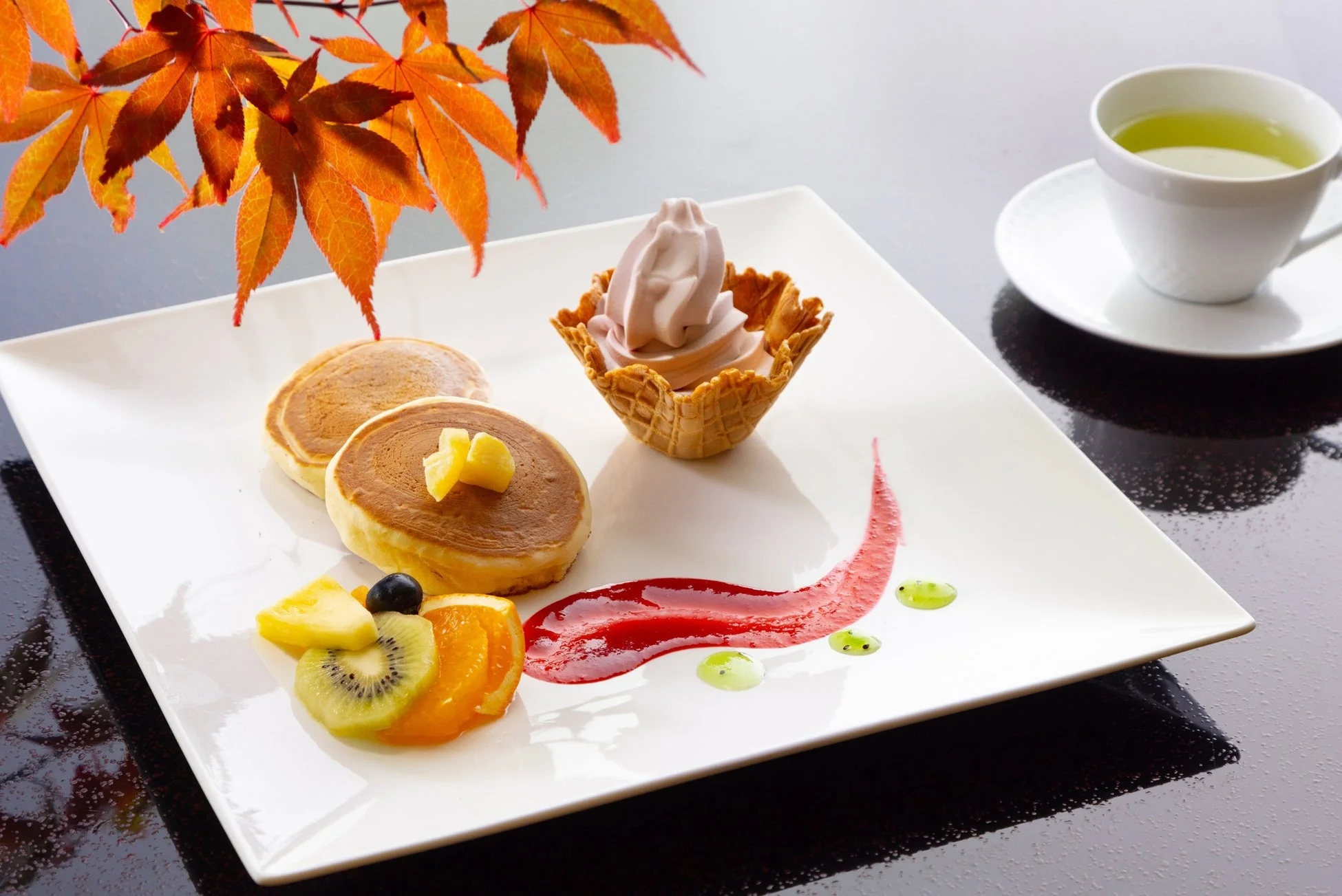 秋の紅葉を背景に、パンケーキ、ソフトクリーム、カットフルーツが美しく盛り付けられたデザートプレートと、温かいお茶
