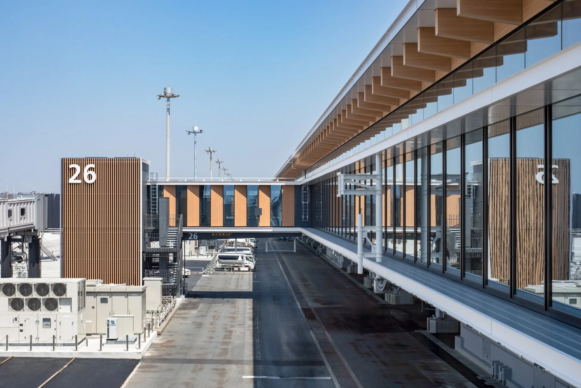 青空の下、近代的な空港ターミナルビルと搭乗橋が写っています。ガラス張りの建物には木製の庇があり、ゲート26の表示が見えます。エプロンには車両や設備が配置されています。