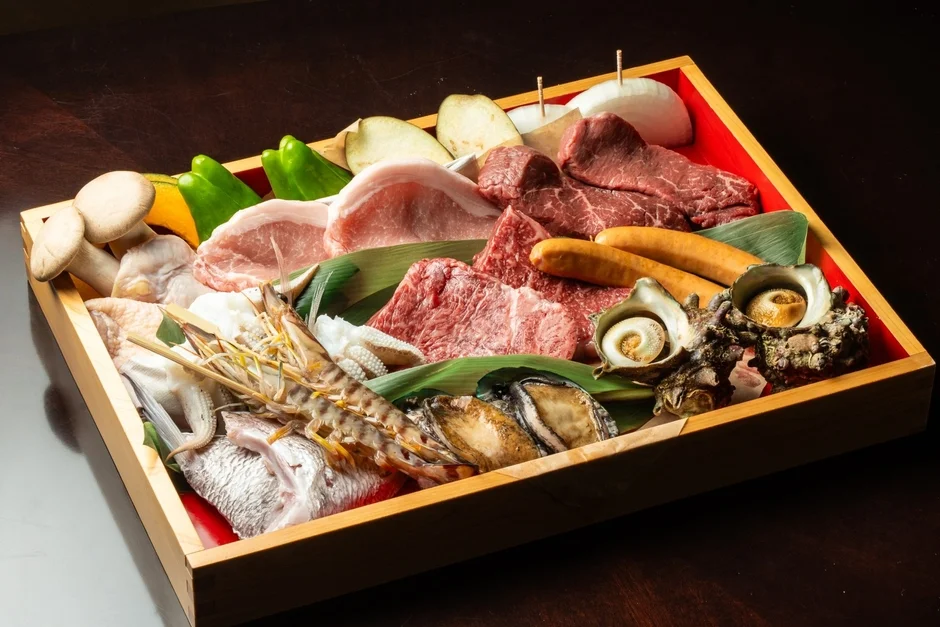 木製の箱に、牛肉、豚肉、ソーセージ、海老、魚、アワビ、サザエ、様々な野菜が美しく盛り付けられた豪華なバーベキュー用食材セット。新鮮さが際立つ。