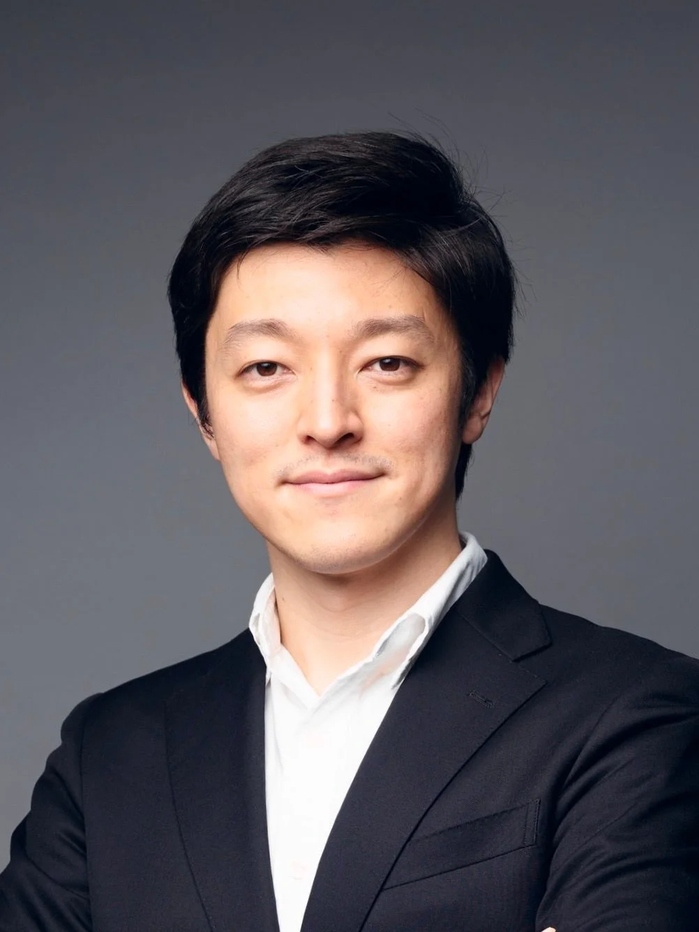 グレーの背景の前で、黒いスーツと白いシャツを着用したアジア人男性のポートレート。カメラに視線を向け、穏やかな笑顔を浮かべている。