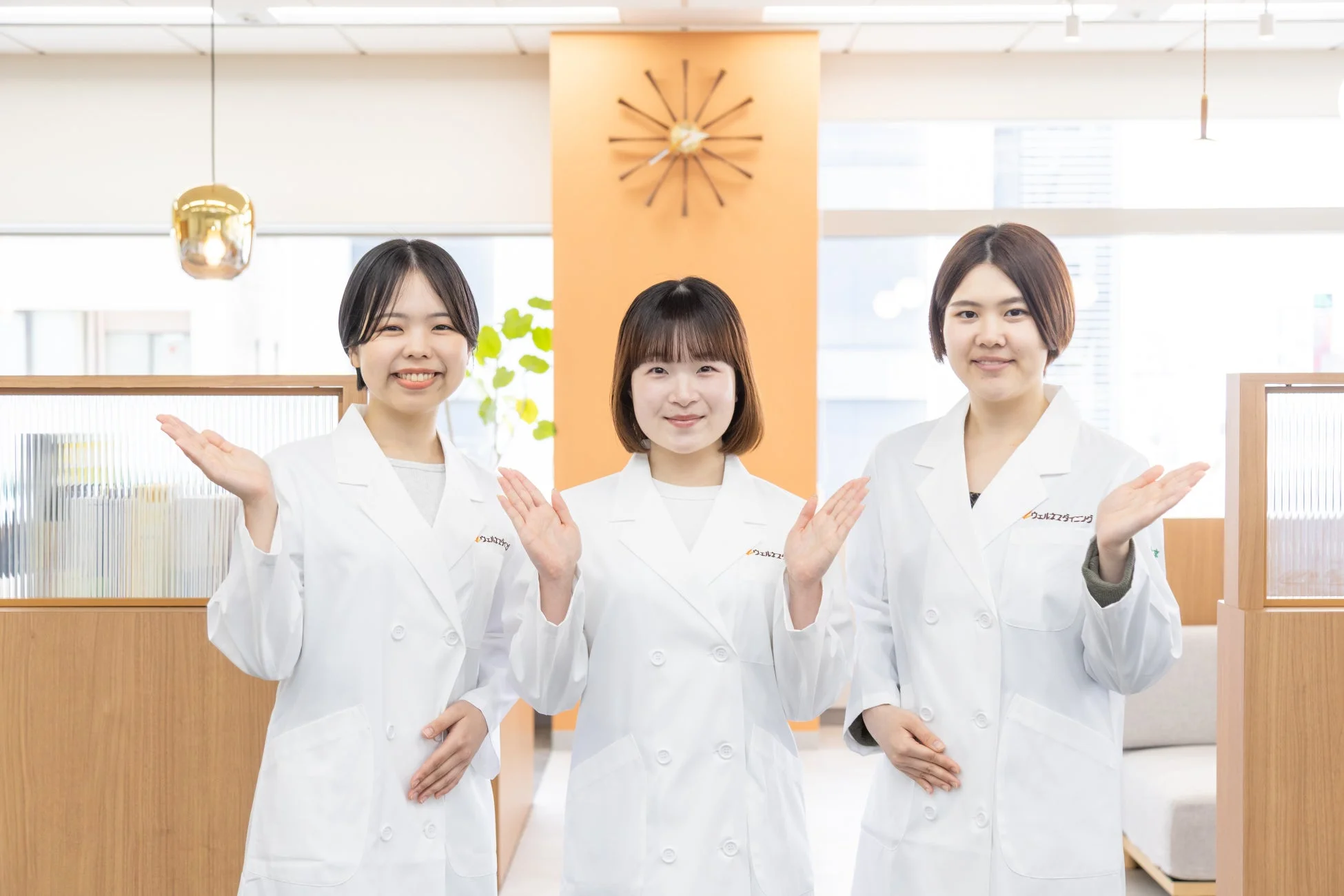 白衣を着た3人の女性が笑顔で両手を広げ、何かを案内している様子