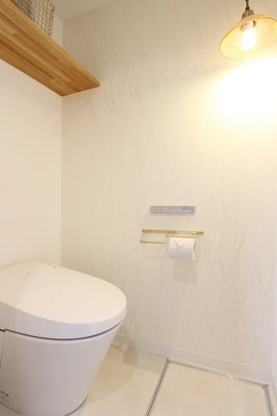 明るく清潔感のあるトイレ空間です。白いモダンな便器、ゴールドのトイレットペーパーホルダーが設置され、上部には木製の棚があります。波打つような模様の白い壁紙と暖色系の照明が落ち着いた雰囲気を醸し出しています。