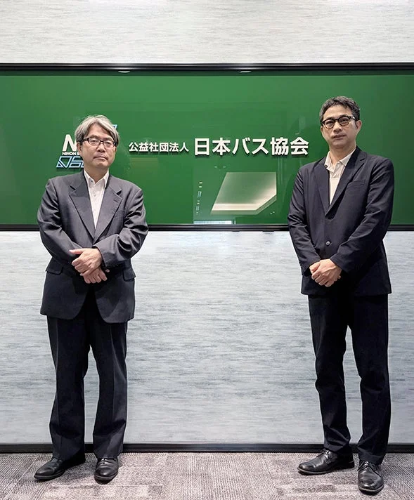 公益社団法人日本バス協会の看板を背景に、スーツ姿の男性2人が並んで立っているビジネスシーンの写真です。会議室のような場所で撮影されており、真剣な表情でカメラを見ています。