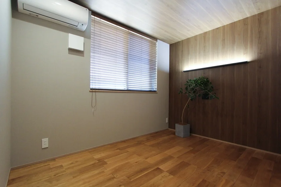 木目のフローリングとウッドパネルの壁が印象的なモダンな部屋です。窓にはブラインドがあり、壁にはエアコンと横長の照明、鉢植えの観葉植物が配置されています。シンプルで落ち着いた雰囲気の内装が特徴です。
