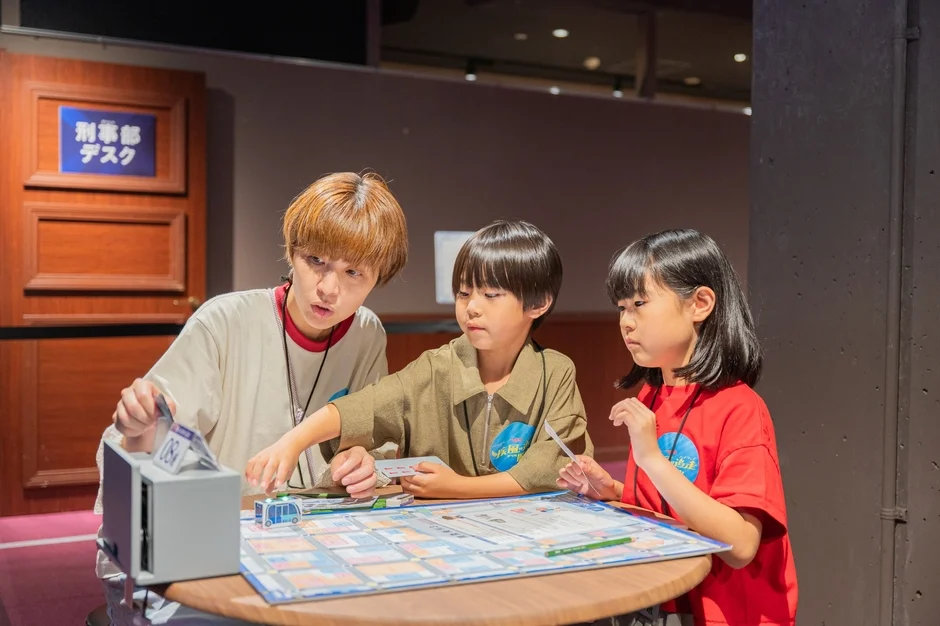 刑事部デスクと書かれた場所で、大人が2人の子供にボードゲームのような体験型アクティビティを教えている様子。