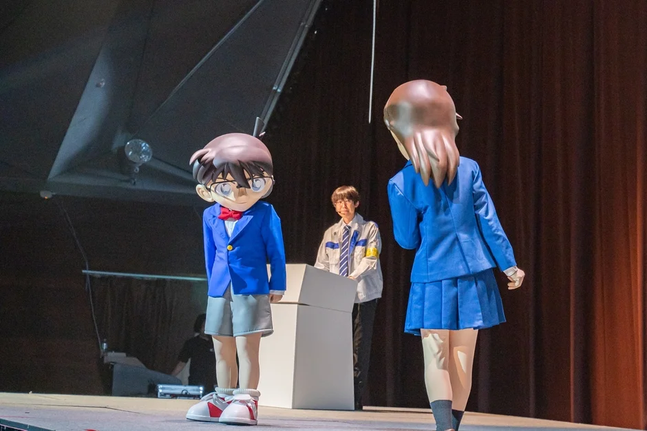 「名探偵コナン」の江戸川コナンと、青い制服を着た女性キャラクターの着ぐるみがステージ上にいます。