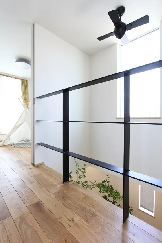 白い壁と木目のフローリングが特徴のモダンな住宅の室内。黒いアイアン手すりのある吹き抜けからは緑が見え、奥にはティピーテントがある。シーリングファンも設置された開放的な空間。