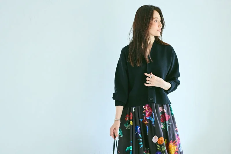 明るい背景の前で、黒いカーディガンとカラフルな花柄スカートを合わせた女性が横を向いているポートレート。