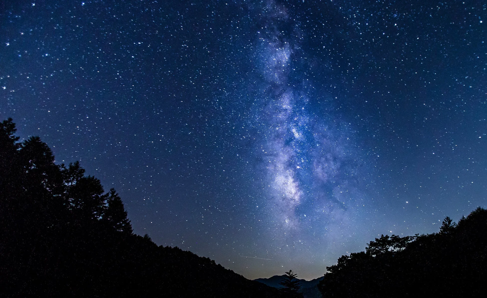 満点の星が輝く夜空に、雄大な天の川が鮮やかに浮かび上がっています。下部には山や木々のシルエットが広がり、神秘的で美しい自然の風景を捉えた一枚です。