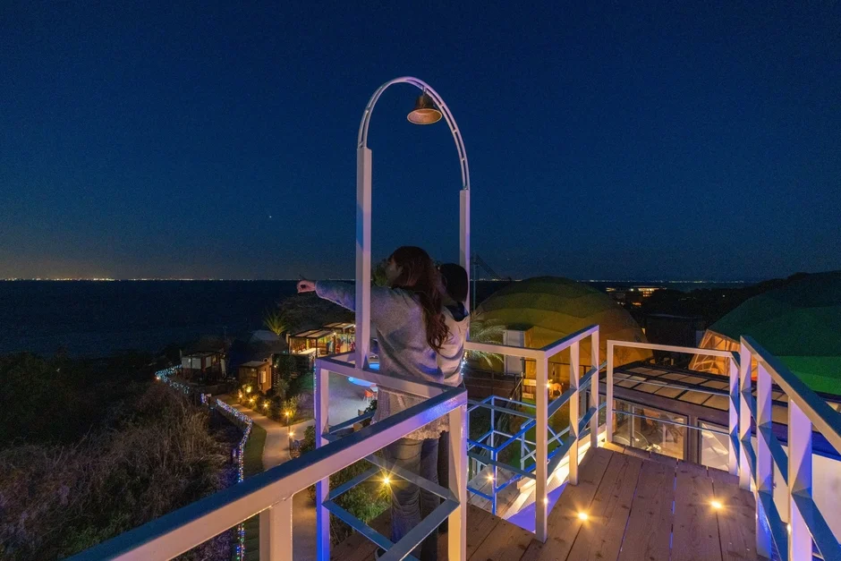 夜の展望台から、カップルが遠くの海の向こうに広がる街の夜景と星空を眺めている。ライトアップされた施設が幻想的な雰囲気を醸し出している。
