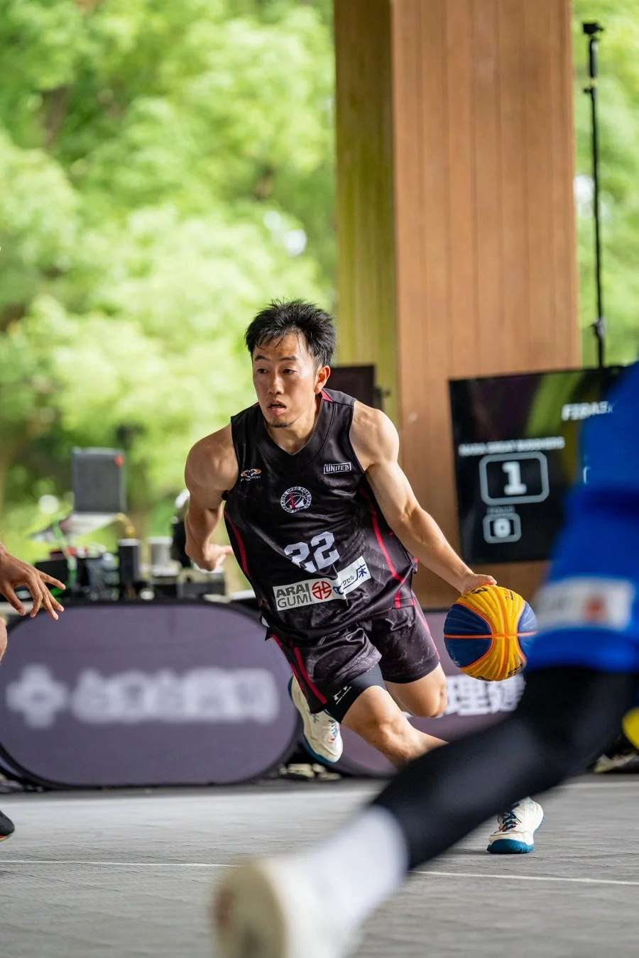黒いユニフォームを着た男性バスケットボール選手が、屋外コートで黄色と青のボールをドリブルしている様子。
