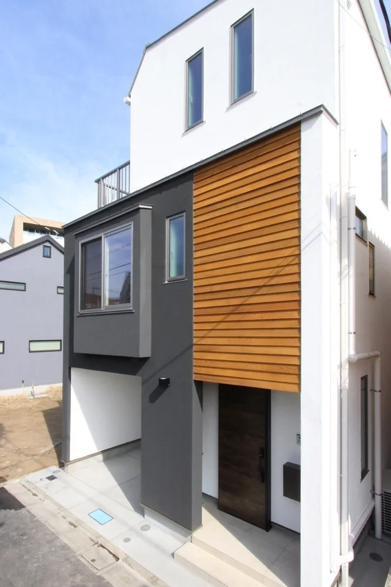 青空の下に建つ、白、チャコールグレー、木目調のパネルが特徴的な3階建ての現代的なデザイン住宅の外観です。1階部分にはガレージスペースが見られます。