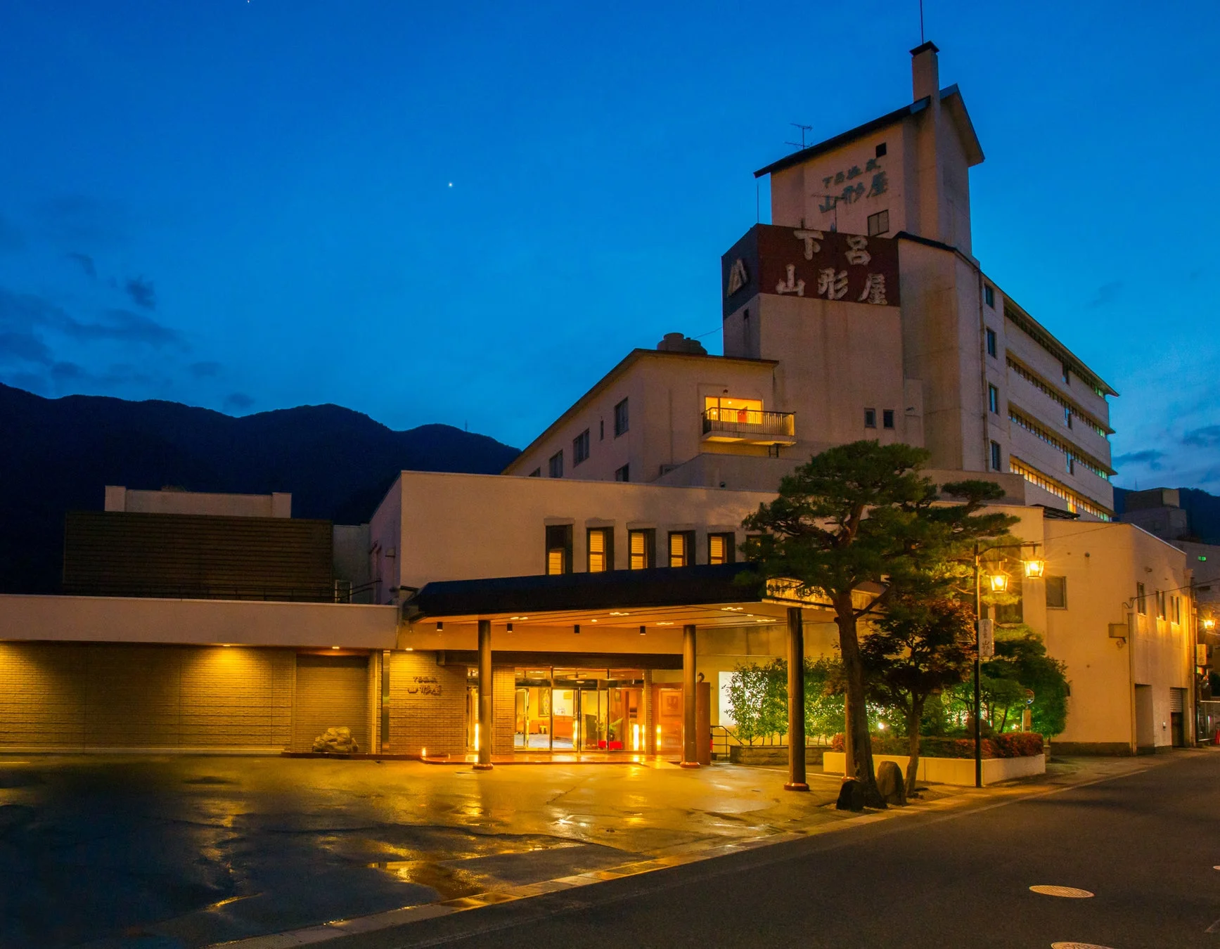 下呂温泉にある「山形屋」という旅館の夜景です。青い空と山の背景に、温かくライトアップされた建物の入り口が印象的で、地面には光が反射しています。