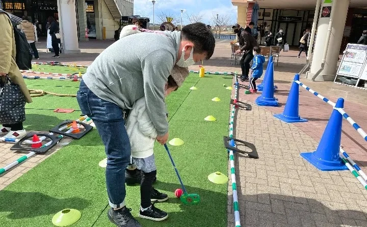 屋外で、大人が子供にミニゴルフのようなパッティングゲームを教えている様子です。人工芝のコースにはコーンが並び、親子で楽しんでいる様子が伺えます。背景には商業施設が見えます。
