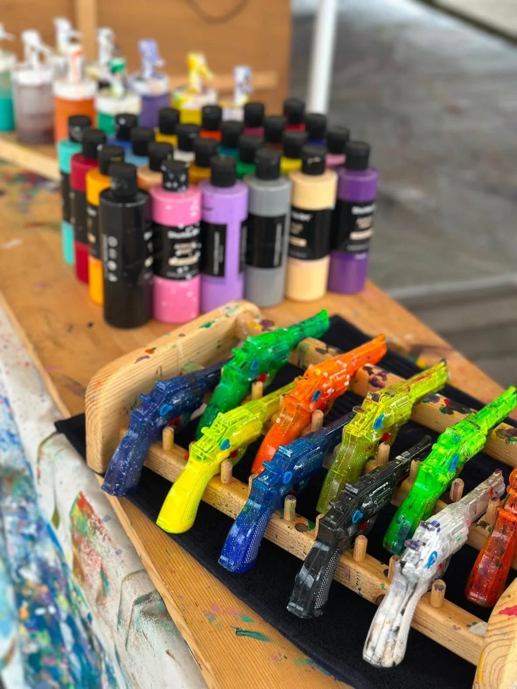背景に様々な色の絵の具ボトルが多数並び、手前にはカラフルなスケルトンのおもちゃの銃が木製ラックに飾られている。絵の具が飛び散った作業台が、創造的な活動の場であることを示唆している。