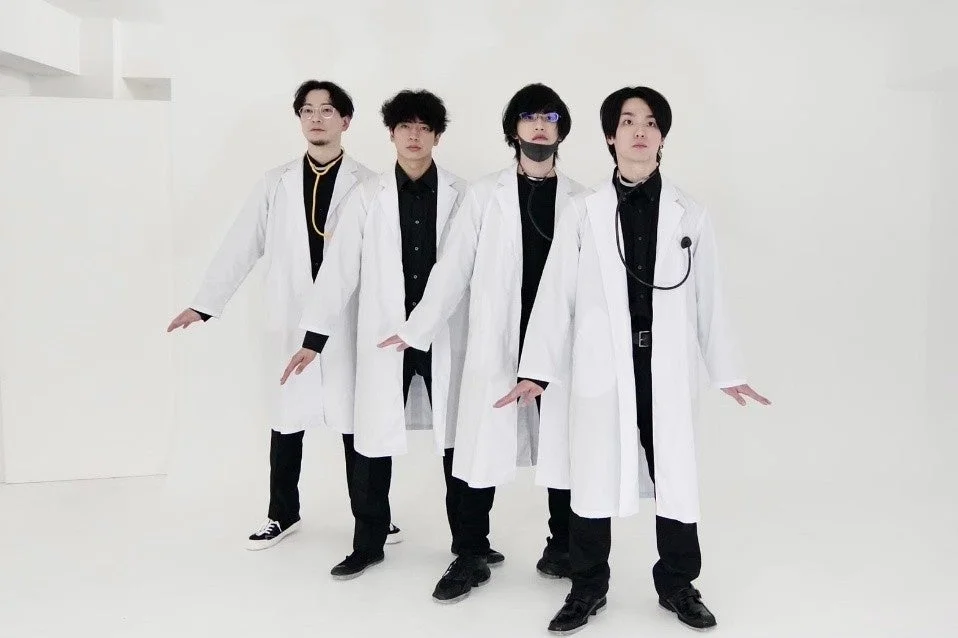 白いスタジオで、白衣と黒い服装に身を包んだ4人の男性が立っています。数人は聴診器を首にかけ、全員が特徴的なポーズをとっており、清潔感とプロフェッショナルな雰囲気が漂うグループショットです。