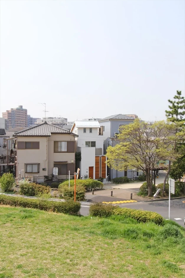 日本の住宅街の風景を捉えた画像。手前には緑豊かな草地があり、奥には伝統的な家屋と現代的なデザインの白い家が並んでいます。