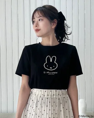 ミッフィーのイラストが描かれた黒いTシャツとドット柄のプリーツスカートを着用した女性が、白い壁を背景に立っています。髪はリボンでポニーテールにまとめています。