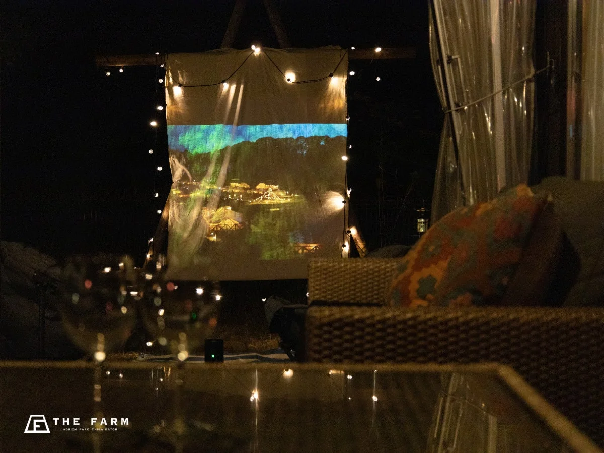 夜のアウトドア空間で、布に映し出された映像が光るプロジェクターと、周囲を飾るイルミネーションが特徴的な画像です。手前にはソファとグラスがあり、リラックスした雰囲気の中で映像鑑賞が楽しめる様子が伺えます。
