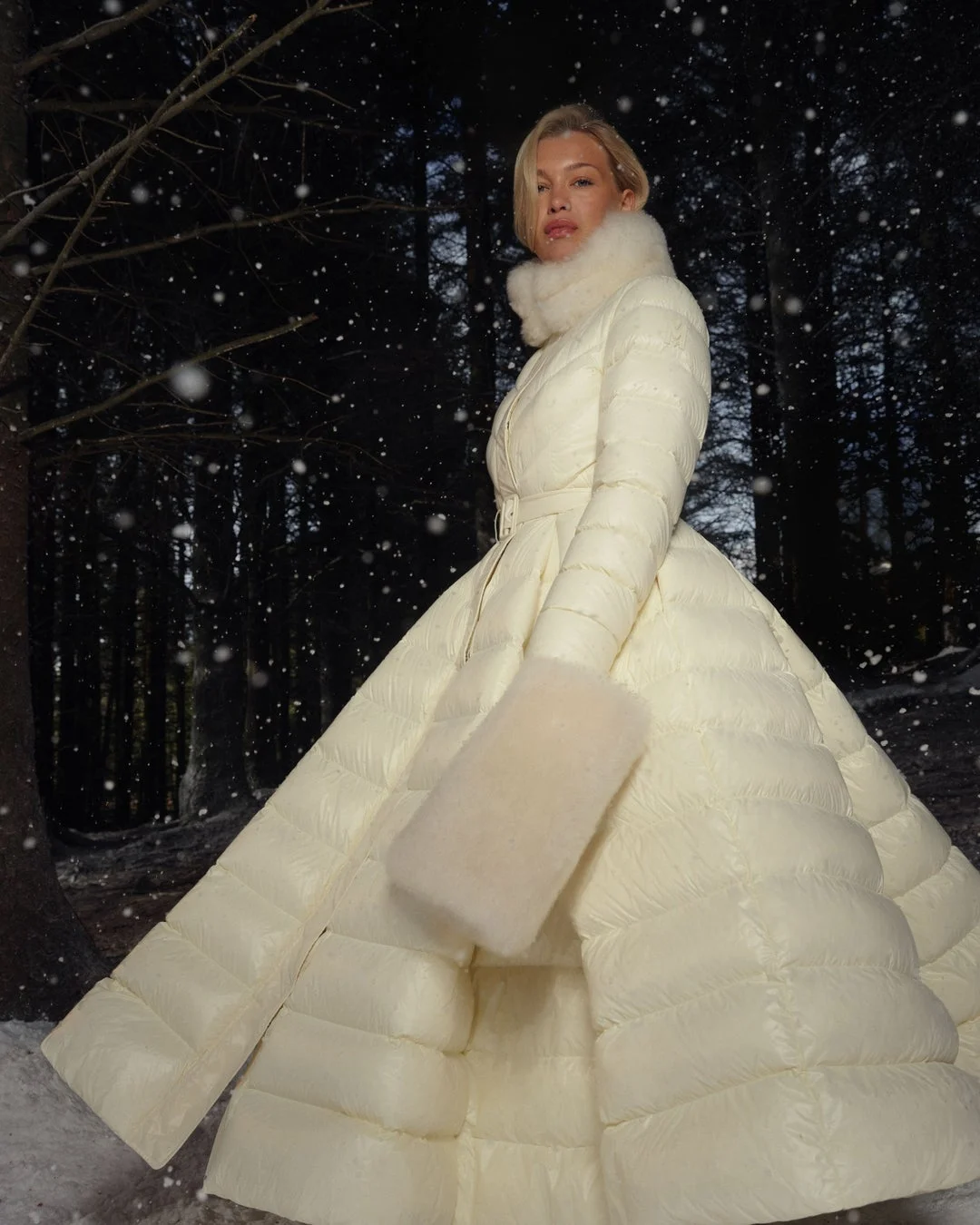 雪が降る夜の森で、白いキルティングのロングコートとファーをまとった女性のファッションポートレート。冬の幻想的な雰囲気を捉えています。