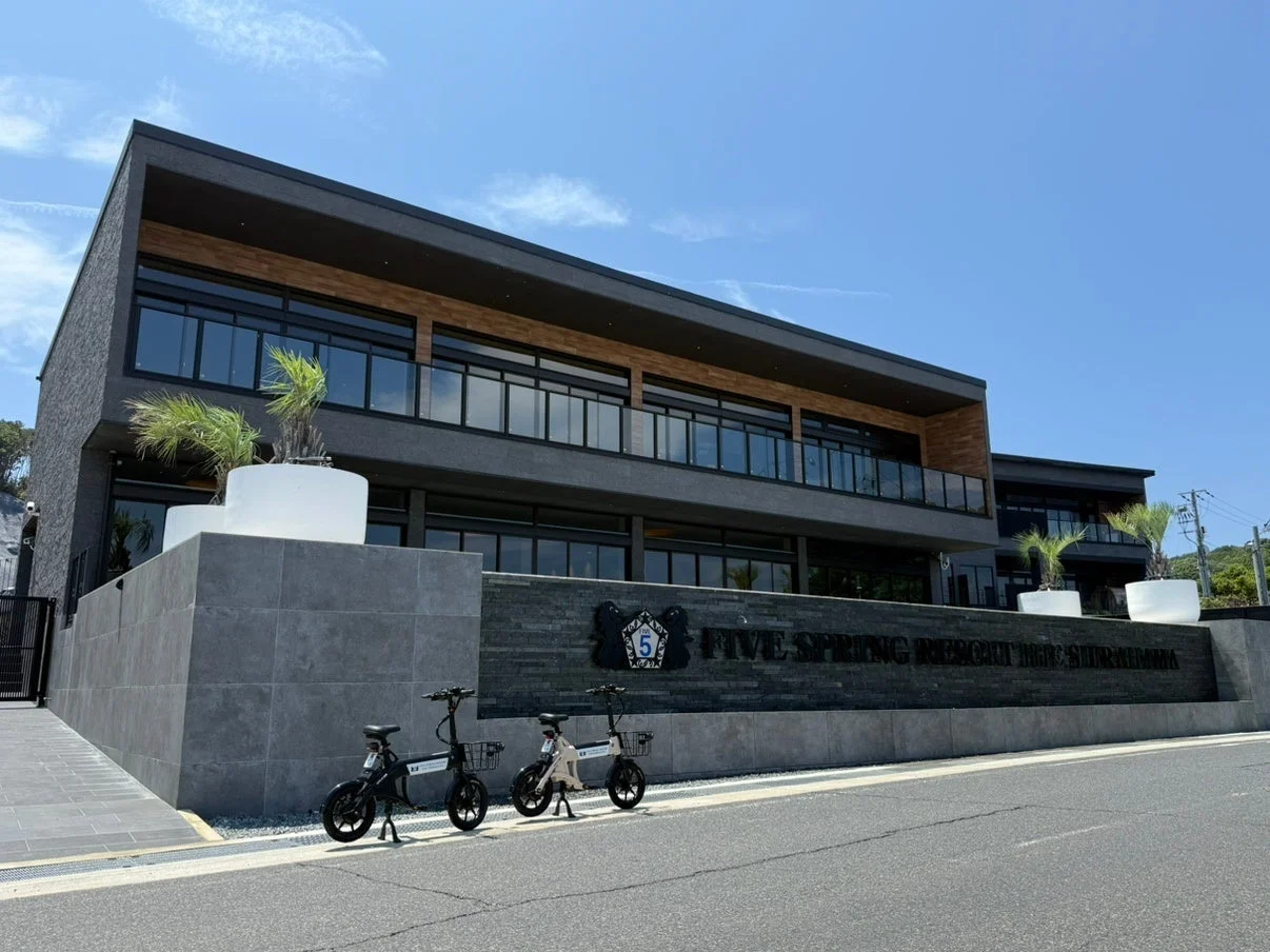 青空の下、モダンな外観のホテル「FIVE SPRING RESORT HOTEL SHIRAHAMA」が写っています。建物の前には2台の電動自転車が停められており、リゾート感のある雰囲気が漂っています。