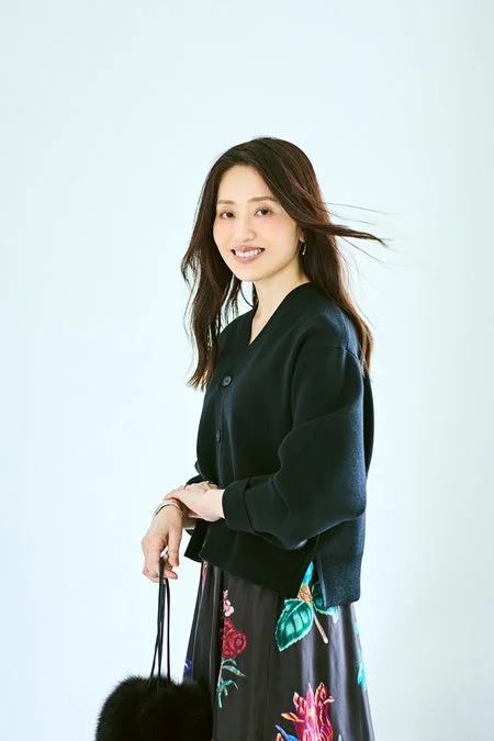 明るい背景のスタジオで微笑むアジア人女性のポートレート。黒のカーディガンとカラフルな花柄ロングスカートを着用し、黒いファーのバッグを持っている。