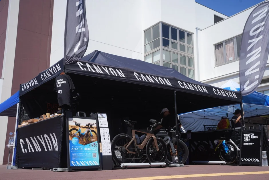 CANYONブランドの自転車試乗会イベントが屋外で開催されている様子です。黒いテントの下には複数のロードバイクが展示され、参加者が試乗できるようになっています。関連グッズや情報も提供されており、賑やかな雰囲気の中で自転車愛好家が集まっています。