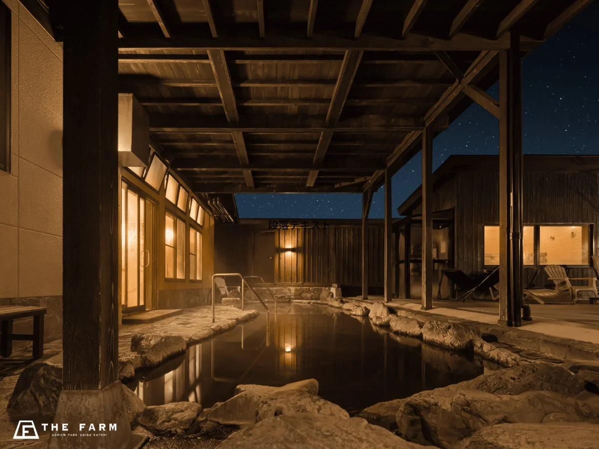 夜空の下、湯気が立ち上る露天風呂の情景です。木造の屋根と温かい光に包まれた石造りの浴槽が、星空と共に穏やかなリラックス空間を演出しています。