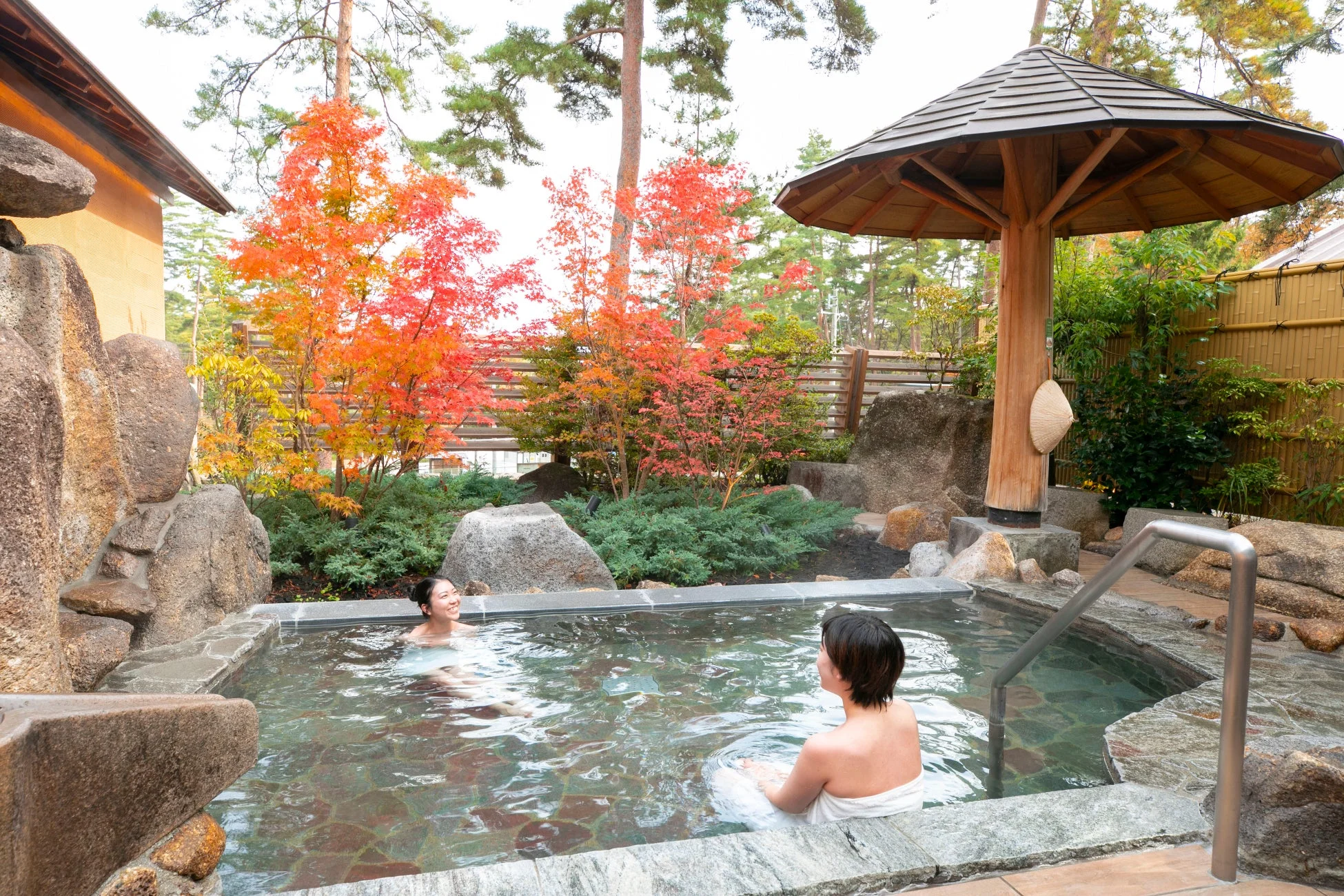 紅葉が美しい秋の露天風呂で、二人の女性がリラックスして温泉を楽しんでいます。岩や緑に囲まれた自然豊かな空間で、癒しのひとときを過ごしている様子が伝わります。