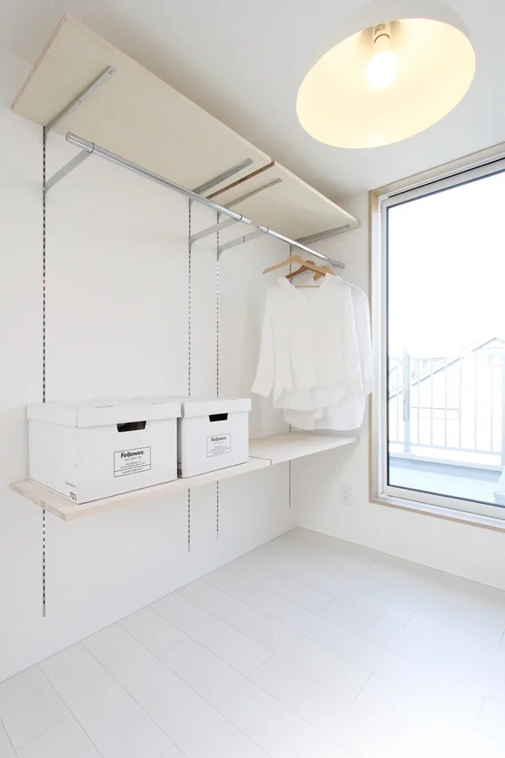 白を基調とした明るい部屋で、壁面には可動式の棚とハンガーラックが設置されています。収納ボックスと白いブラウスが置かれ、シンプルで整理された空間が広がっています。