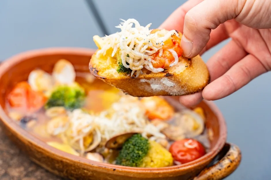 温かいアヒージョをバゲットにのせて食べる様子を捉えた写真です。しらすとエビがたっぷり乗ったバゲットを、アサリやブロッコリー、ミニトマトなどが入ったアヒージョの器から取り上げており、食欲をそそる一品です。