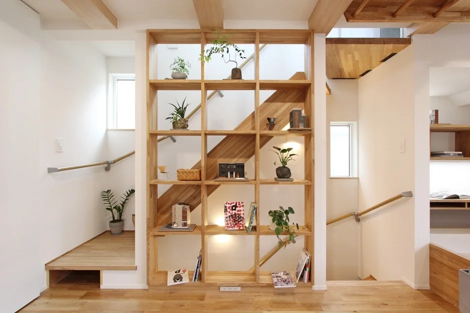 明るい木材と白い壁が特徴のモダンな住宅のインテリア。中央には観葉植物や本が飾られた大きな格子状の木製棚があり、空間を区切りつつ階段へと繋がる開放的なデザインが目を引きます。暖かく整理された居住空間を演出しています。