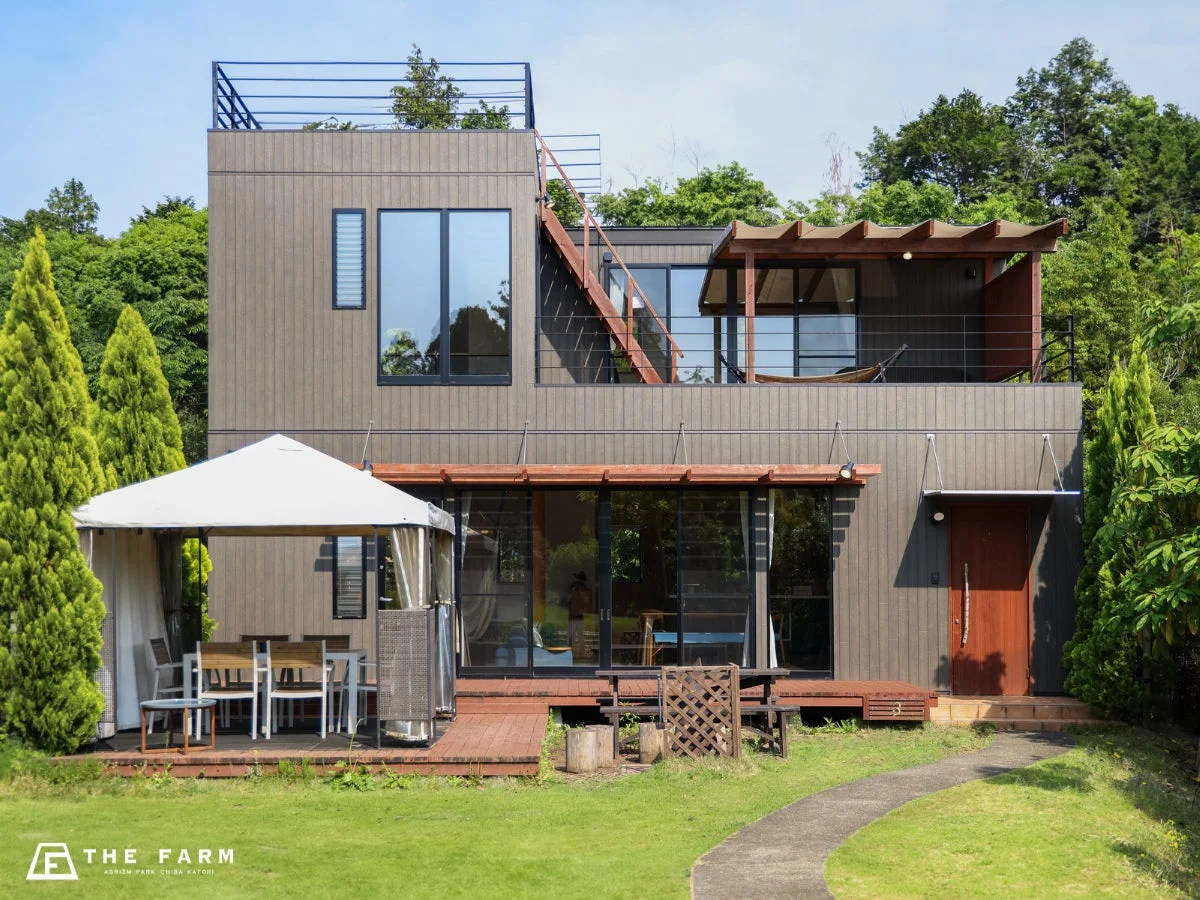 自然豊かな環境に建つモダンな2階建ての建物です。ウッドデッキには屋外ダイニング用のテントやハンモックが設置されており、ゆったりとした時間を過ごせるリゾートのような雰囲気が特徴です。
