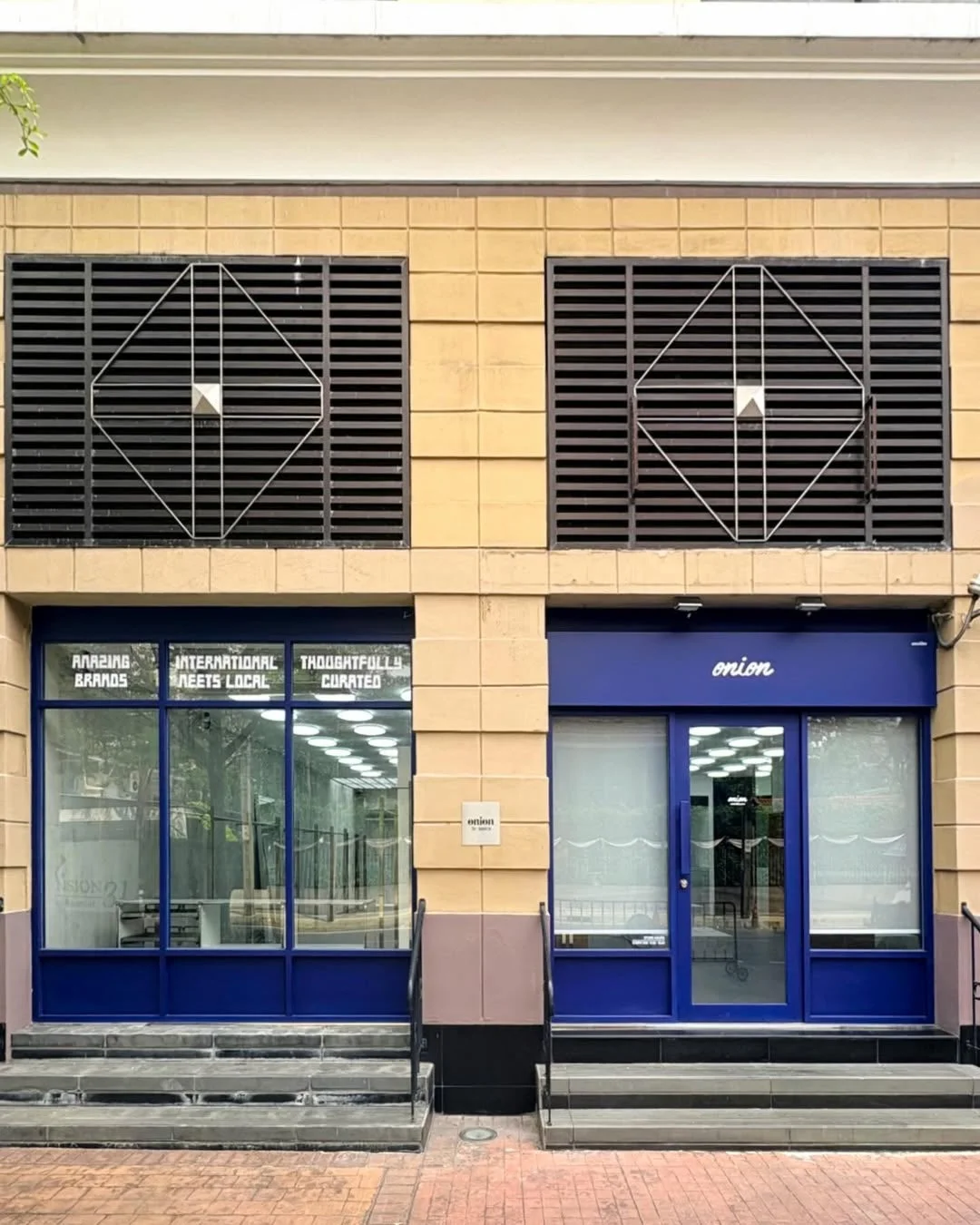 2つの店舗が並ぶ建物の正面を捉えた写真。左の店舗の窓には「AMAZING BRANDS」などのメッセージがあり、右の店舗は「onion」という店名が青い扉の上に表示されている。上部には格子状のルーバーが特徴的なデザインの建物です。