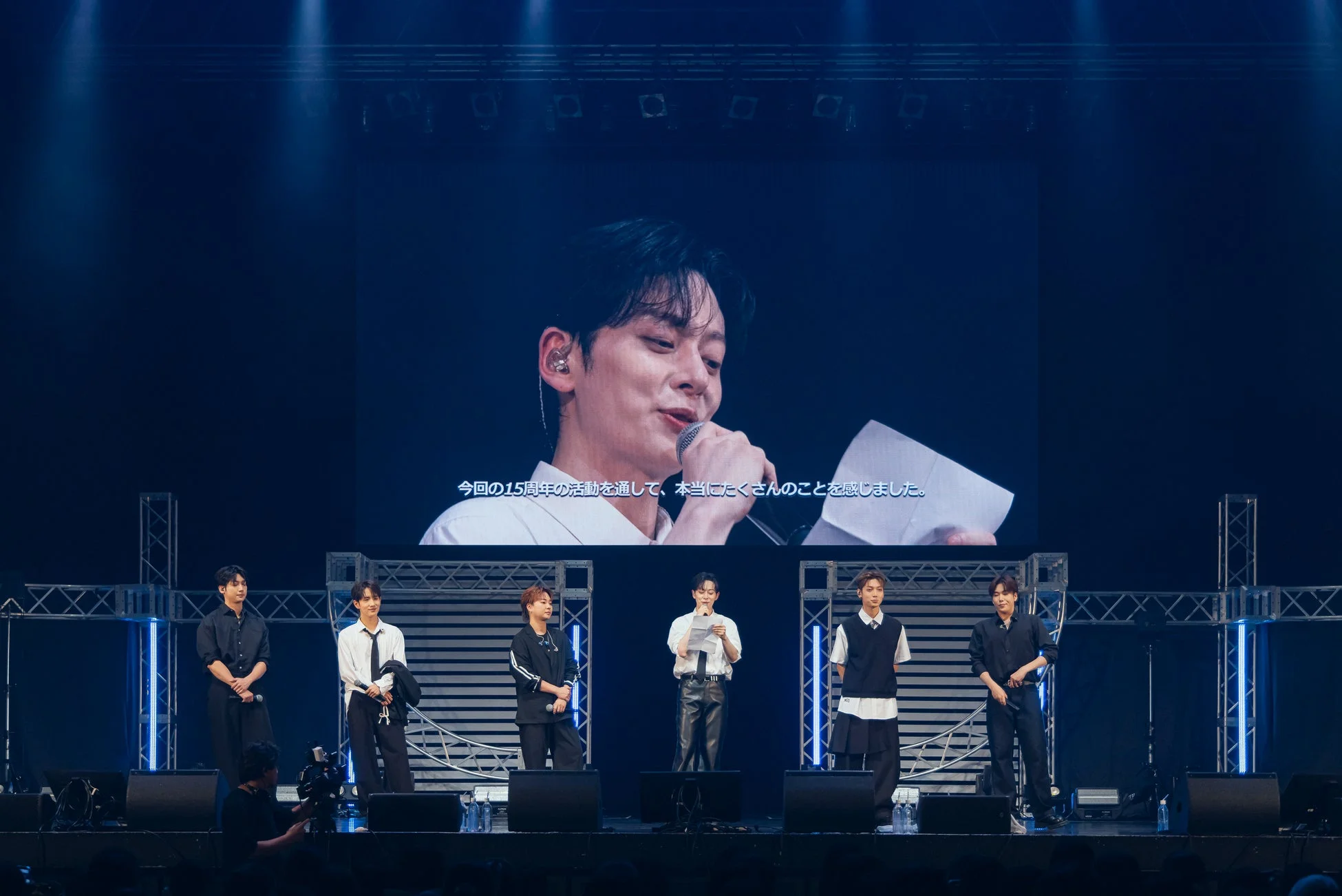 ステージ上で6人組の男性グループがイベントを行っており、背後の大型スクリーンにはメンバーの一人がマイクを持って話す姿と「今回の15周年の活動を通して、本当にたくさんのことを感じました。」というメッセージが映し出されています。