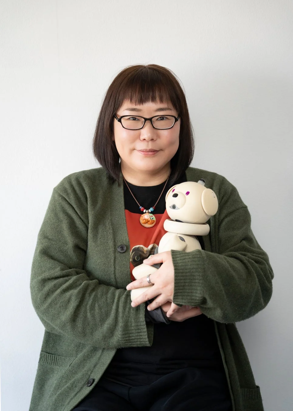 緑のカーディガンを着て眼鏡をかけた女性が、白い背景の前でクリーム色の犬型ロボット(AIBO)を抱きかかえているポートレート