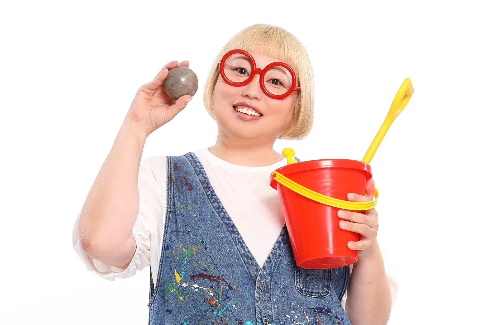 金髪ボブで赤い丸眼鏡をかけた女性が、笑顔でカメラを見つめています。片手には灰色の玉、もう片方には赤いバケツとスコップを持ち、ペンキが飛び散ったデニムのオーバーオールを着ています。楽しげな雰囲気のスタジオ撮影です。