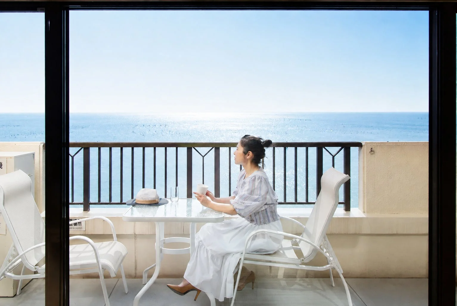 女性がホテルのバルコニーで、広大な海を眺めながらカップを片手にくつろいでいる