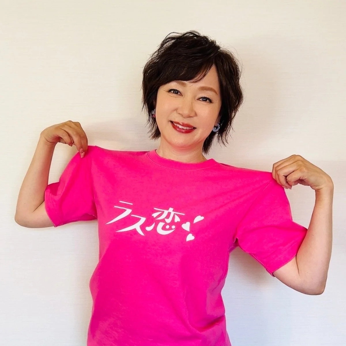 笑顔の女性が、白い壁を背景に「ラス恋!」と書かれた鮮やかなピンクのTシャツを着ています。ショートヘアで明るい表情をしています。