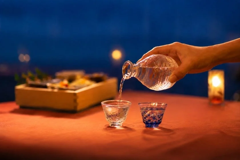 夜の雰囲気の中、手が透明な徳利から小さなグラスに日本酒を注いでいる