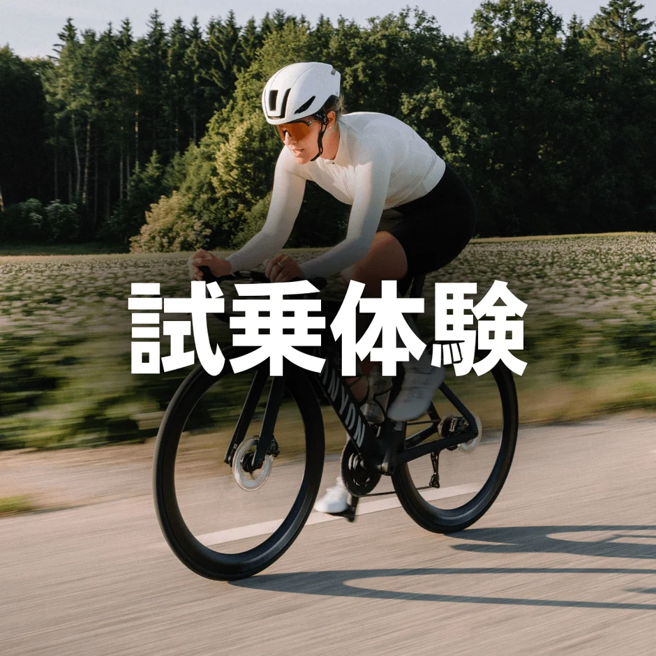 白いヘルメットとサングラスを着用した人がロードバイクに乗って舗装された道を走行しています。背景には木々や畑が広がり、画像には「試乗体験」という文字が重ねられています。