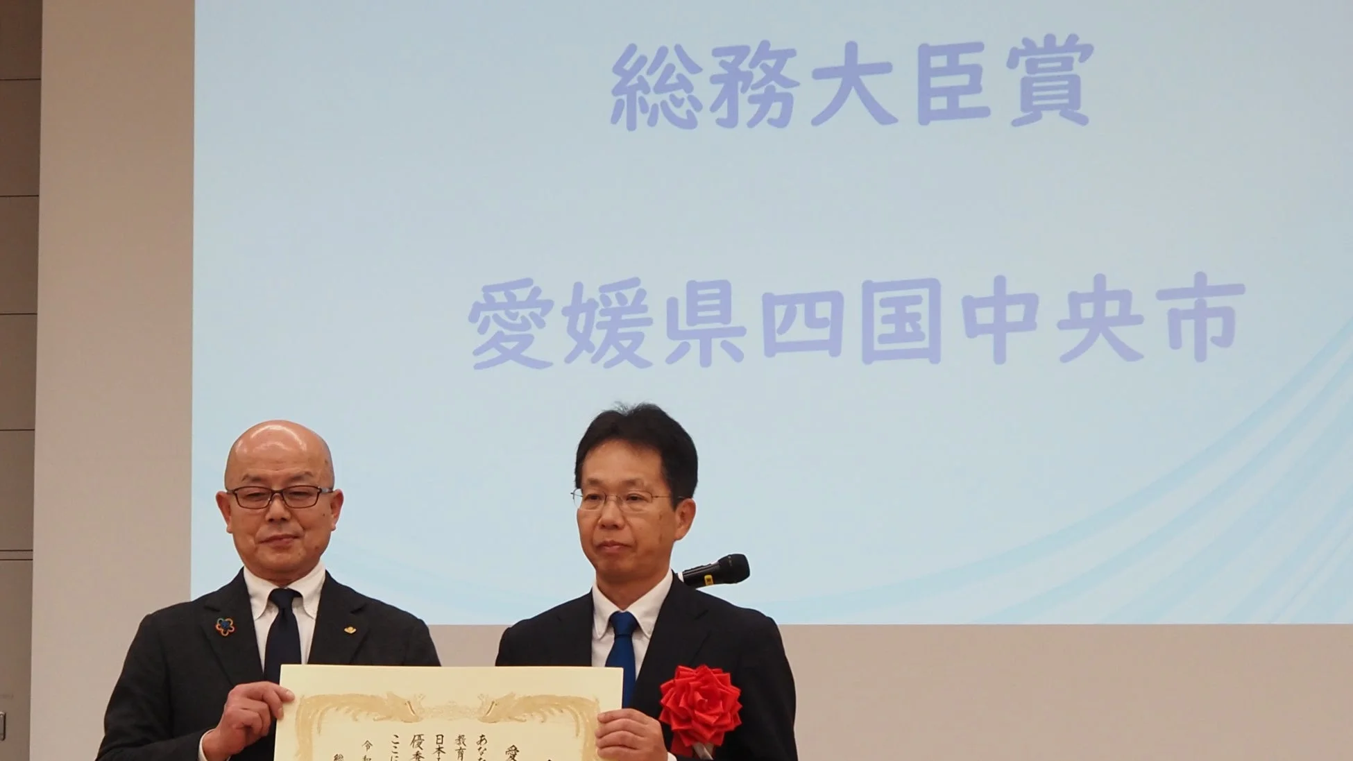 総務大臣賞を受賞した愛媛県四国中央市の関係者が、表彰式で賞状を手にしている様子。壇上のスクリーンには受賞内容と自治体名が示されています。