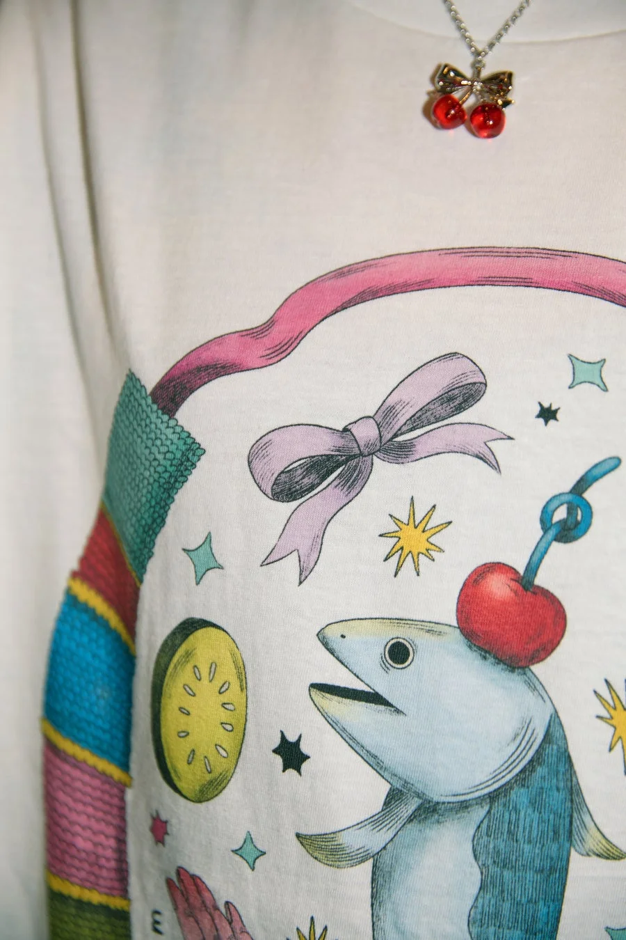 白いTシャツに、チェリーを頭に乗せた魚やリボン、星、黄色い果物のスライスなど、カラフルでファンタジックなイラストが描かれています。上部にはチェリーのネックレスも着用されています。